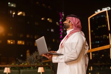 رجل سعودي يحمل كمبيوتر محمول في شرفة ليلية