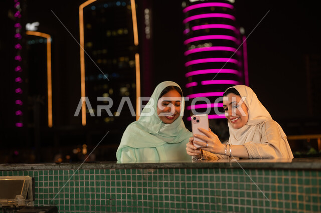 نساء عربيات يستخدمن الهاتف أمام أفق المدينة ليلاً