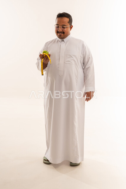 رجل سعودي مع تفاحة وشريط قياس على خلفية بيضاء