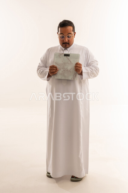 رجل سعودي يعاني من السمنة يمسك ميزان وزن في الاستوديو