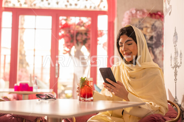 امرأة عربية ترتدي شيلة بيج تستخدم الهاتف في مقهى