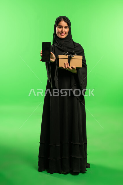امرأة سعودية بالعباءة مع هدية وهاتف بخلفية خضراء