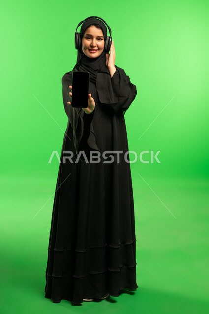 امرأة سعودية ترتدي سماعات وتمسك هاتف ذكي أمام كروما خضراء