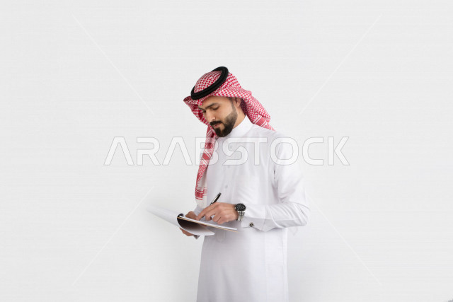بورتريه رجل أعمال عربي سعودي يقف و يكتب في مجلد ، ورق ملاحظات ، خلفية بيضاء