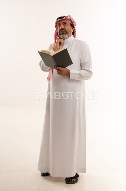 رجل سعودي بالزي التقليدي يفكر ممسكاً بكتاب في استوديو