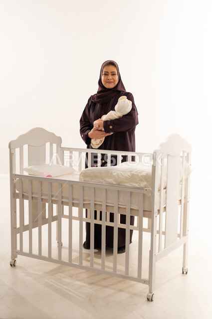 Saudi Woman Holding Swaddled Infant Behind White Crib