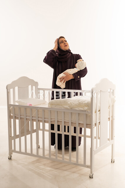 Exhausted Saudi Mother Holding Newborn Near White Crib