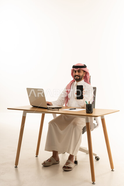 رجل أعمال سعودي يمسك هاتف ذكي أمام كمبيوتر محمول