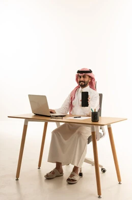 رجل أعمال سعودي يمسك هاتف ذكي أمام كمبيوتر محمول