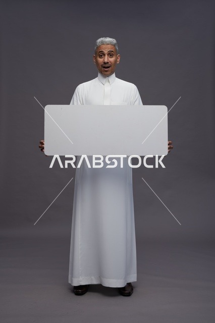 رجل سعودي يحمل لوحة بيضاء فارغة بتعبير وجه مندهش في استوديو