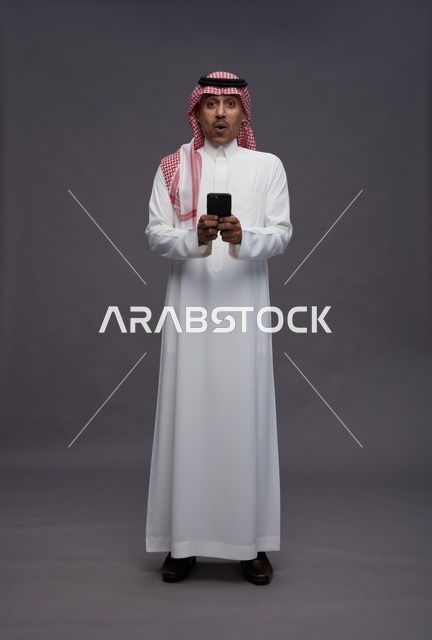 رجل سعودي مندهش يمسك هاتفاً ذكياً بخلفية رمادية