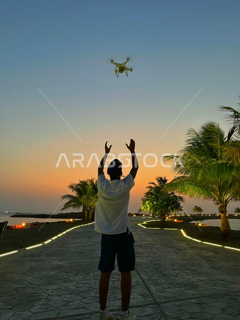 صورة من الخلف لشاب عربي خليجي سعودي، الاستعداد للإمساك بطائرة بدون طيار، طائرة درون، غروب الشمس، طائرة تصوير جوي
