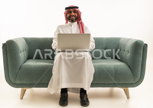 رجل سعودي مع حاسوب محمول يجلس على أريكة خلفية بيضاء