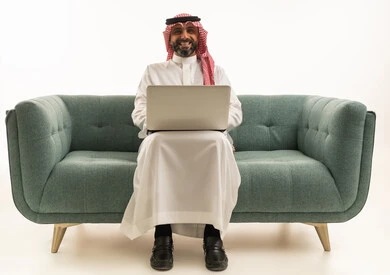 رجل سعودي مع حاسوب محمول يجلس على أريكة خلفية بيضاء