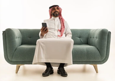 Saudi Man in Traditional Thobe Holding Smartphone on Sofa