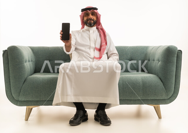 Saudi Man in Traditional Thobe Holding Smartphone on Sofa