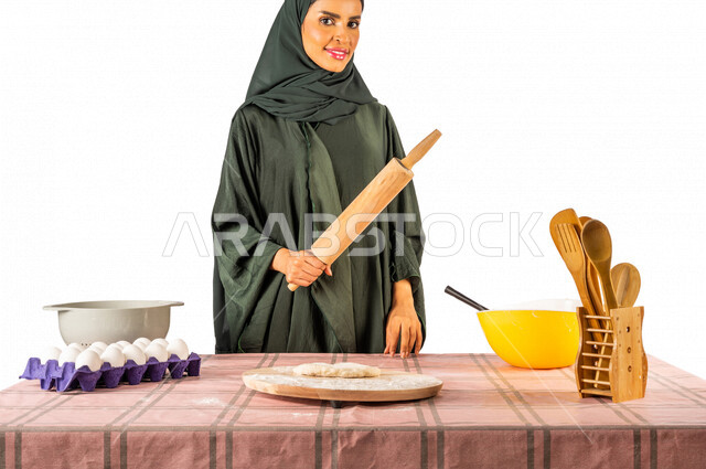 بورتريه بصورة مقربة لمرأة خليجية سعودية، تقف أمام طاولة عليها مكونات خاصة بالعجين وأواني الخبز وأدوات خاصة بالمطبخ والعجين، تحمل بيدها أداة فرد العجين اليد، رقاقة العجين، الشوبك (النشابة)، مفهوم إعداد المخبوزات والمعجنات الشهية، تحضير الطعام، ربة منزل