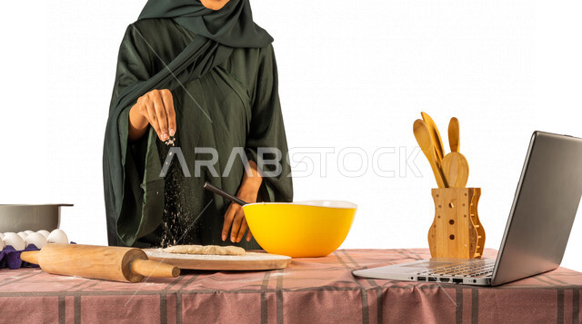 بورتريه لمرأة خليجية سعودية، تقف أمام طاولة عليها مكونات خاصة بالعجين وأواني الخبز وأدوات خاصة بالمطبخ والعجين، إعداد المخبوزات الشهية، إستخدام جهاز الحاسوب المحمول، بث مباشر، مشاركة تحضير الوصفات على مواقع التواصل الاجتماعي، إستخدام أجهزة تقنية حديثة