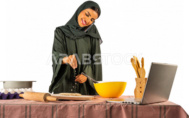 Portrait of a Saudi Gulf woman, standing in front of a table with ingredients for dough and baking utensils, preparing delicious baked goods, making a mobile phone call, using a laptop, broadcasting live, sharing recipes on social media, communicating wit