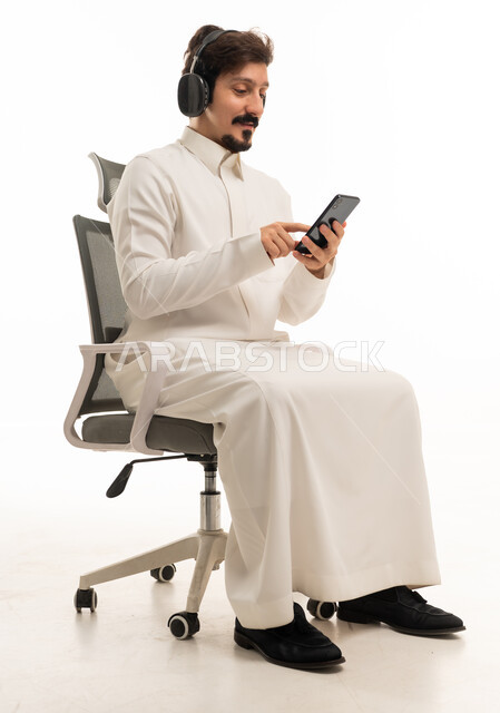 رجل سعودي يستخدم الهاتف الذكي وسماعات الرأس على كرسي مكتب