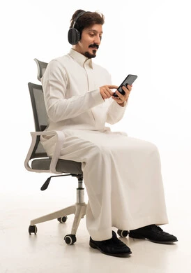 رجل سعودي يستخدم الهاتف الذكي وسماعات الرأس على كرسي مكتب