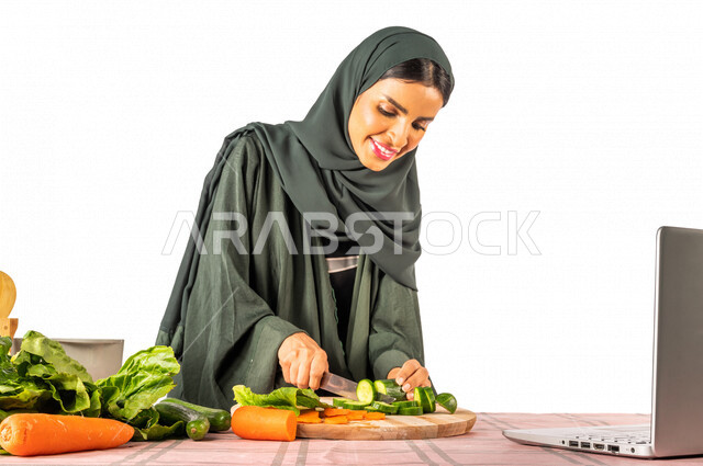 Portrait of a Saudi Gulf woman, standing in front of a table with a group of fresh vegetables, making a video call via a laptop, sharing preparing recipes on social media, preparing salads and healthy dishes, the concept of a healthy diet, live broadcast