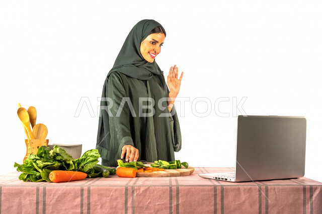 Portrait of a Saudi Gulf woman, standing in front of a table with a group of fresh vegetables, making a video call via a laptop, sharing preparing recipes on social media, preparing salads and healthy dishes, the concept of a healthy diet, live broadcast