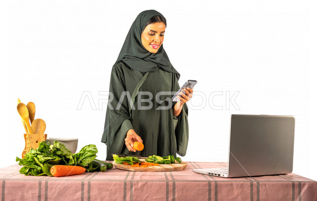 Portrait of a Saudi Gulf woman, standing in front of a table with a group of fresh vegetables, using her mobile and laptop, sharing preparing recipes on social media, preparing healthy dishes, surfing the Internet, using modern technology devices