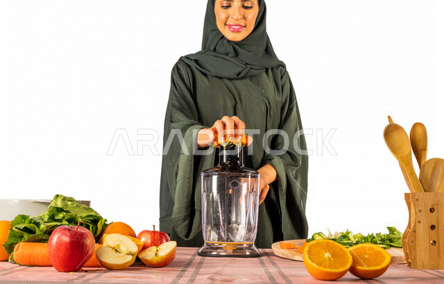 بورتريه بصورة مقربة لمرأة خليجية سعودية، مجموعة من مكونات الفواكه الطازجة على الطاولة لتحضير العصير، تقوم بعصر البرتقال بإستخدام آلة العصارة، أجهزة كهرومنزلية، إعداد العصائر الصحية، مفهوم النظام الغذائي الصحي، خلفية بيضاء، ربة منزل ، التدبير المنزلي