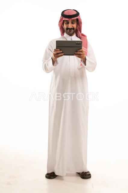 رجل سعودي بالثوب والشماغ يستخدم جهاز لوحي خلفية بيضاء