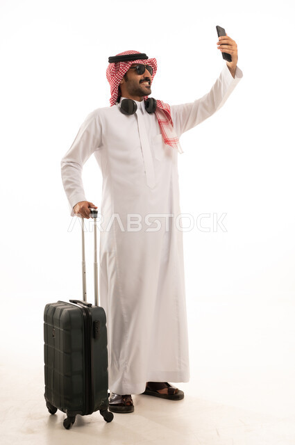 توثيق اللحظات السعيدة عبر الهاتف المحمول, استخدام جهاز تقني حديث ومتطور, بورتريه لرجل عربي سعودي خليجي يرتدي الزي الثوب الابيض والشماغ يضع نظارة شمسية ويقوم بالتقاط صورة السيلفي التذكارية عبر الجوال يمسك بيدة شنطة سفر, الجسم بالطول الكامل, خلفية بيضاء .