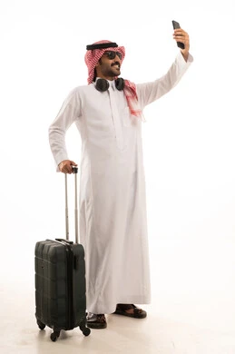 توثيق اللحظات السعيدة عبر الهاتف المحمول, استخدام جهاز تقني حديث ومتطور, بورتريه لرجل عربي سعودي خليجي يرتدي الزي الثوب الابيض والشماغ يضع نظارة شمسية ويقوم بالتقاط صورة السيلفي التذكارية عبر الجوال يمسك بيدة شنطة سفر, الجسم بالطول الكامل, خلفية بيضاء .