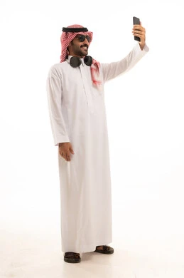 توثيق اللحظات السعيدة عبر الهاتف المحمول, استخدام جهاز تقني حديث ومتطور, بورتريه لرجل عربي سعودي خليجي يرتدي الزي الثوب الابيض والشماغ يضع نظارة شمسية ويقوم بالتقاط صورة السيلفي التذكارية عبر الجوال, الجسم بالطول الكامل, خلفية بيضاء .