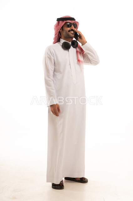 توثيق اللحظات السعيدة عبر الهاتف المحمول, استخدام جهاز تقني حديث ومتطور, بورتريه لرجل عربي سعودي خليجي يرتدي الزي الثوب الابيض والشماغ يضع نظارة شمسية ويقوم بالتقاط صورة السيلفي التذكارية عبر الجوال, الجسم بالطول الكامل, خلفية بيضاء .