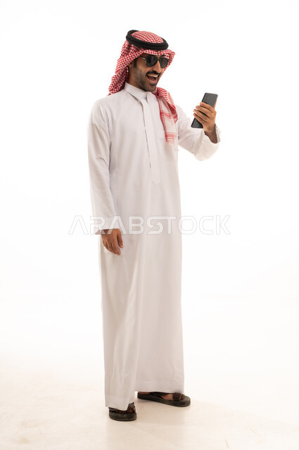 تصفح مواقع التواصل الاجتماعي, استخدام جهاز تقني حديث ومتطور,  بورتريه لرجل عربي سعودي خليجي يرتدي الزي الثوب الابيض والشماغ يضع نظارة شمسية ويقوم باستخدام الهاتف المحمول, الجسم بالطول الكامل, خلفية بيضاء .