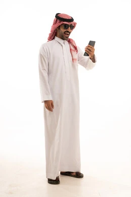 تصفح مواقع التواصل الاجتماعي, استخدام جهاز تقني حديث ومتطور,  بورتريه لرجل عربي سعودي خليجي يرتدي الزي الثوب الابيض والشماغ يضع نظارة شمسية ويقوم باستخدام الهاتف المحمول, الجسم بالطول الكامل, خلفية بيضاء .