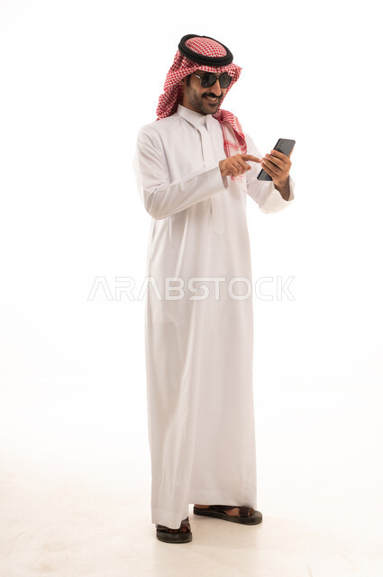 تصفح مواقع التواصل الاجتماعي, استخدام جهاز تقني حديث ومتطور,  بورتريه لرجل عربي سعودي خليجي يرتدي الزي الثوب الابيض والشماغ يضع نظارة شمسية ويقوم باستخدام الهاتف المحمول, الجسم بالطول الكامل, خلفية بيضاء .