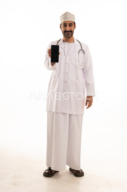 دمج التقنية والتكنولوجيا في الطب والرعاية الصحية، العمل في القطاع الطبي في سلطنة عمان، بورتريه لطبيب عربي خليجي عماني يرتدي الدشداشة والكمة والزي الخاص بالمهنة يضع السماعة الطبية يستخدم  الهاتف المحمول بتعابير السرور، الجسم بالطول الكامل، خلفية بيضاء