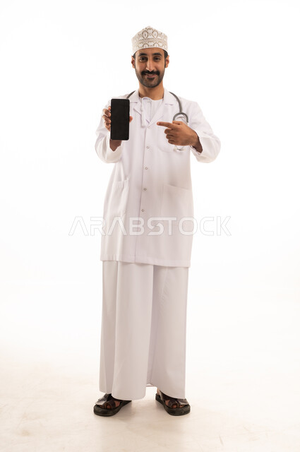 دمج التقنية والتكنولوجيا في الطب والرعاية الصحية، العمل في القطاع الطبي في سلطنة عمان، بورتريه لطبيب عربي خليجي عماني يرتدي الدشداشة والكمة والزي الخاص بالمهنة يضع السماعة الطبية يستخدم  الهاتف المحمول بتعابير السرور، الجسم بالطول الكامل، خلفية بيضاء
