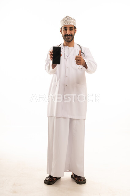 تعابير الموافقة والرضي، دمج التقنية والتكنولوجيا في الطب والرعاية الصحية، العمل في القطاع الطبي في سلطنة عمان، بورتريه لطبيب عربي خليجي عماني يرتدي الدشداشة والكمة والزي الخاص بالمهنة يضع السماعة الطبية يستخدم  الهاتف المحمول ، الجسم بالطول الكامل، خلفية بيضاء