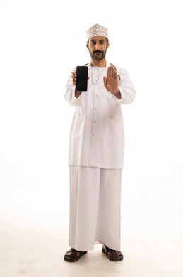 تعابير التوقف عن فعل شئ ما ، دمج التقنية والتكنولوجيا في الطب والرعاية الصحية، العمل في القطاع الطبي في سلطنة عمان، بورتريه لطبيب عربي خليجي عماني يرتدي الدشداشة والكمة والزي الخاص بالمهنة يضع السماعة الطبية يستخدم  الهاتف المحمول ، الجسم بالطول الكامل، خلفية بيضاء