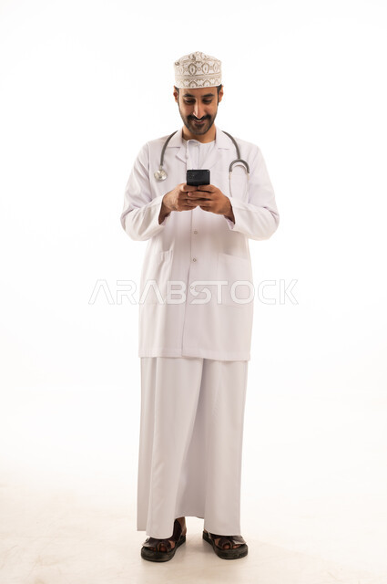 دمج التقنية والتكنولوجيا في الطب والرعاية الصحية، العمل في القطاع الطبي في سلطنة عمان، بورتريه لطبيب عربي خليجي عماني يرتدي الدشداشة والكمة والزي الخاص بالمهنة يضع السماعة الطبية يستخدم  الهاتف المحمول بتعابير السرور، الجسم بالطول الكامل، خلفية بيضاء