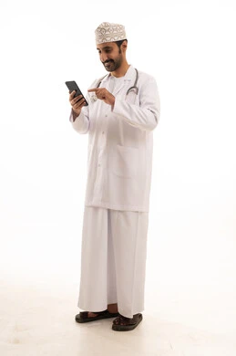 دمج التقنية والتكنولوجيا في الطب والرعاية الصحية، العمل في القطاع الطبي في سلطنة عمان، بورتريه لطبيب عربي خليجي عماني يرتدي الدشداشة والكمة والزي الخاص بالمهنة يضع السماعة الطبية يستخدم  الهاتف المحمول بتعابير السرور، الجسم بالطول الكامل، خلفية بيضاء
