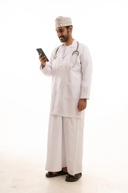 دمج التقنية والتكنولوجيا في الطب والرعاية الصحية، العمل في القطاع الطبي في سلطنة عمان، بورتريه لطبيب عربي خليجي عماني يرتدي الدشداشة والكمة والزي الخاص بالمهنة يضع السماعة الطبية يستخدم  الهاتف المحمول بتعابير السرور، الجسم بالطول الكامل، خلفية بيضاء