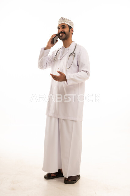 دمج التقنية والتكنولوجيا في الطب والرعاية الصحية، العمل في القطاع الطبي في سلطنة عمان، بورتريه لطبيب عربي خليجي عماني يرتدي الدشداشة والكمة والزي الخاص بالمهنة يضع السماعة الطبية يستخدم  الهاتف المحمول بتعابير السرور، الجسم بالطول الكامل، خلفية بيضاء
