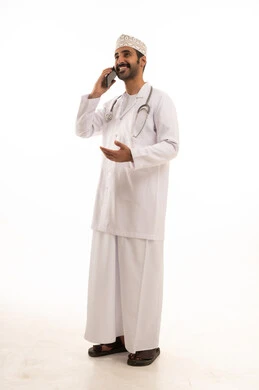 دمج التقنية والتكنولوجيا في الطب والرعاية الصحية، العمل في القطاع الطبي في سلطنة عمان، بورتريه لطبيب عربي خليجي عماني يرتدي الدشداشة والكمة والزي الخاص بالمهنة يضع السماعة الطبية يستخدم  الهاتف المحمول بتعابير السرور، الجسم بالطول الكامل، خلفية بيضاء