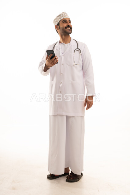 دمج التقنية والتكنولوجيا في الطب والرعاية الصحية، العمل في القطاع الطبي في سلطنة عمان، بورتريه لطبيب عربي خليجي عماني يرتدي الدشداشة والكمة والزي الخاص بالمهنة يضع السماعة الطبية يستخدم  الهاتف المحمول بتعابير السرور، الجسم بالطول الكامل، خلفية بيضاء