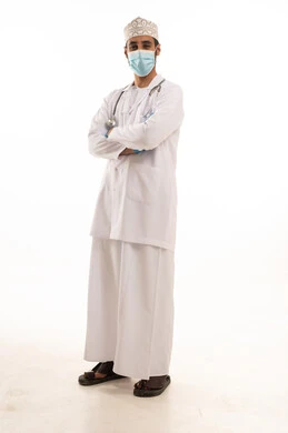 Following safety procedures to prevent the coronavirus, a portrait of an Arab Gulf Omani doctor wearing a dishdasha, turban, medical coat, and mask, holding a stethoscope, working in the health sector, full-body, white background.