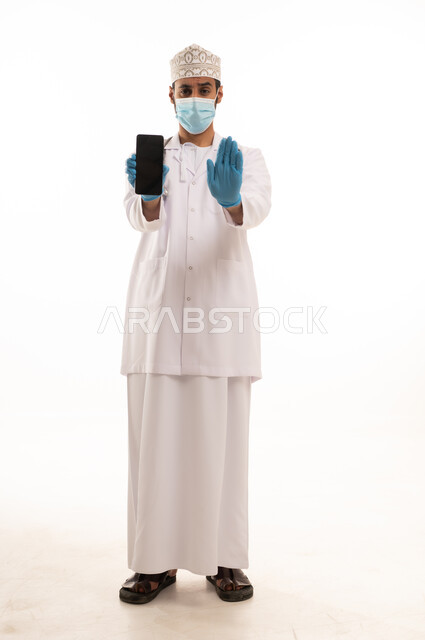 تعابير التوقف عن فعل شئ ما ، دمج التقنية والتكنولوجيا في الطب والرعاية الصحية، العمل في القطاع الطبي في سلطنة عمان، بورتريه لطبيب عربي خليجي عماني يرتدي الدشداشة والكمة والزي الخاص بالمهنة يضع السماعة الطبية يستخدم  الهاتف المحمول ، الجسم بالطول الكامل، خلفية بيضاء