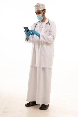 Following safety procedures to prevent the coronavirus, integrating technology and tech in medicine and healthcare, working in the medical sector in the Sultanate of Oman, a portrait of an Arab Gulf Omani doctor wearing a dishdasha and kumma and the professional attire, wearing a stethoscope and using a mobile phone with expressions of joy, full-body, white background.